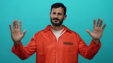 Turuncu hapishane tulumu giyen İspanyol sakallı adam mavi arka planda duruyor. Elleri havada ve sakin bir şekilde teslim ol ya da dur senaryosu öneriyor..