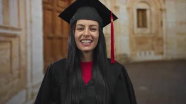 Mezuniyet kepi ve cübbesi takmış genç İspanyol bir kadın tarihi bir arka planla eski bir kasaba sokaklarında neşeyle gülüyor. Eğitimsel başarıyı dışarıda kutluyor..