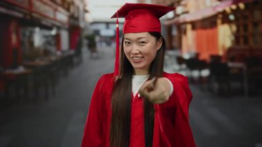 Kırmızı mezuniyet elbisesi giymiş bir kadın şehir terasında bulanık arka planlı bir jest yapıyor..
