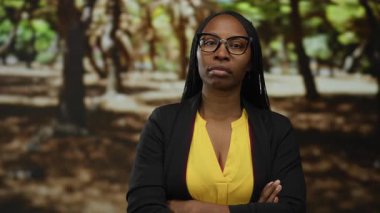 Kıvırcık saçlı bir kadın parkta kolları çapraz duruyor ve ciddi bir ifade takınıyor gözlük ve sarı bluz giyiyor..
