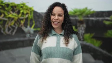 Dışarıda gülümseyen gözlüklü, çizgili süveterli, yeşil çiçekli ve arka planda taş duvarlı bir kadın..