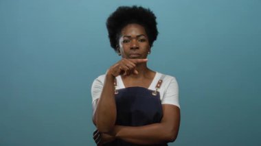 Önlüklü Afrikalı Amerikalı kadın, izole edilmiş mavi arka plana karşı düşünceli duruyor..