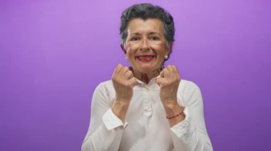 Gri saçlı, canlı pembe bir arka plan önünde heyecanını ifade eden son sınıf öğrencisi bir kadın. Kapalı yumruklar ve parlak bir gülümsemeyle neşe ve pozitiflik sergiliyor..