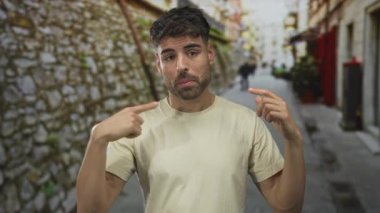 Taş duvar ve bulanık yayalar ile dar bir sokakta göğsü gösteren genç adam parmağı; belirsizlik şüphesi kafa karışıklığı.