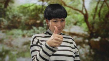 Açık yeşil bir parkta açık elleri olan çizgili süveter giyen genç bir kadın açık havada sunum yapıyor ya da sunum yapıyor..