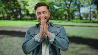 Güneşli bir park ortamında gülümseyen sakallı genç adam, sıradan bir kot ceket giyiyor, açık havada mutluluğu ve rahatlığı ifade ediyor yemyeşil ve ağaçlarla çevrili..