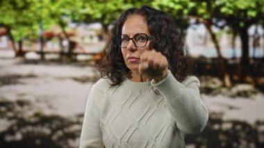 Parkta kıvırcık saçlı İspanyol kadın, gözlük ve süveter takıyor, etrafındakileri dikkatle dinliyor..