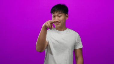 Genç Çinli bir adam, beyaz bir gömlek giyerken izole edilmiş pembe bir arka plan üzerinde neşeli jestler yapıyor, samimi ve esprili bir hava yayıyor..