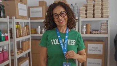 Yardım odasında gülümseyen gönüllü bir üniforma giyen bir kadın. Etrafı yiyecek bağışları ve malzemelerle çevrili ve raflarla çevrili..