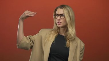 Elleri turuncu arka planda, güven içinde el kol hareketi yapan gözlüklü ve bej ceketli bir kadın iletişim ve ifadeyi resmediyor..