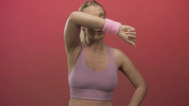 Spor sütyenli bir kadın parlak pembe bir stüdyoda dirseğiyle alnından teri siler. Kararlılık..