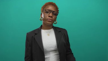 Yeşil bir stüdyo ortamında gözlük ayarlarken alnına dokunan kadın siyah bir ceket giyiyor..