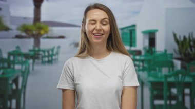 Beyaz gömlekli bir kadın açık hava restoranının terasında yeşil sandalyeli ve masalı bir şekilde gülümsüyor. Rahatlamış, güneşli bir ortam yakalıyor..