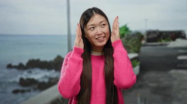 Deniz kenarındaki güvertede gülümseyen uzun saçlı pembe kazak giyen ve arka planda deniz manzaralı sakin ve neşeli bir anı yakalayan bir kadın..