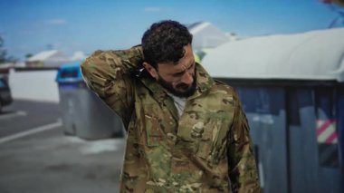 Askeri üniformalı İspanyol bir adam dışarıda çöp konteynırının yanında duruyor. Rahatsızlık içinde boynu ovalıyor, şehir ortamında stresi ya da yorgunluğu gösteriyor..
