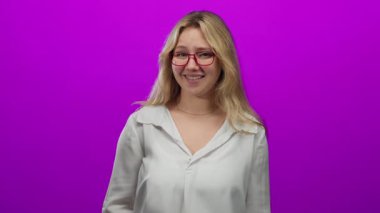 Sarı saçlı ve gözlüklü bir kadın pembe arka plana karşı ezik hareketler yaparak gülüyor..