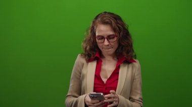 Akıllı telefon kullanan bir kadın, izole edilmiş yeşil arka planda kırmızı bluz ve kırmızı gözlüklü bej ceket giyiyor. Stüdyoda teknoloji ve iletişim kavramlarını tasvir ediyor..