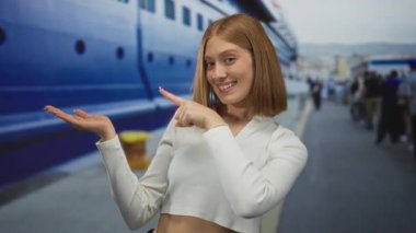 Kadın avuçlarını açık tutuyor ve deniz kıyısındaki boş alanı işaret ediyor; davet karşılama nişanı.