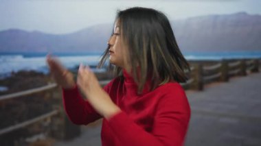 Kırmızılı kadın arka planda okyanus ve dağlarla dolu bir deniz kenarında bağırarak dinamik bir açık hava sahnesi yaratıyor..