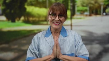 Hispanic senior woman in uniform meditating with hands together in a park, representing peacefulness and dedication outdoors.