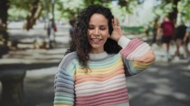 Renkli kazaklı kıvırcık saçlı kadın, güneşli parkta ağaçların bulanık arka planıyla ve yürüyen insanlarla ilgili jest yapıyor..