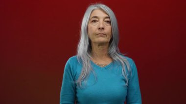 Gri saçlı yaşlı bir kadın canlı kırmızı bir arka plana karşı güvenle duruyor, bilgeliğini ve zarafetini basit turkuaz kıyafetleriyle sergiliyor, izole ve odaklanmış..