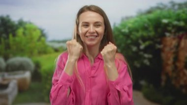 Rahat, doğal bir bahçede gülümseyen, canlı pembe bir gömlek giyen, huzurlu, yemyeşil bir ortamda neşe ve mutluluk gösteren bir kadın..