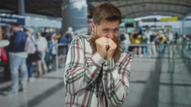 Genç beyaz adam kalabalık havaalanı terminalinde kuyrukta beklerken ağzını yumrukla kapatıyor..