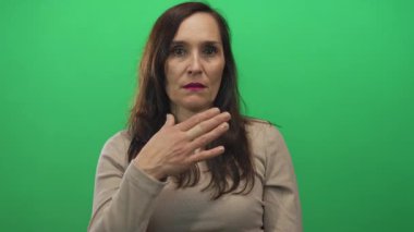 Uzun saçlı bir kadın, elleri göğsünde canlı yeşil bir altyapıya sahip, günlük bej bluz giyiyor, duygularını izole bir ortamda ifade ediyor..