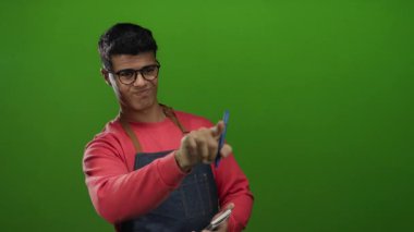 Gözlüklü ve önlüklü genç bir İspanyol, yeşil bir arka plana karşı not alıyor. Profesyonel ve odaklanmış bir garsonun hareket halindeki davranışını yakalıyor..