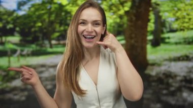 Çağrı jesti olan ve yemyeşil park ortamında gülümseyen parmağıyla işaret eden kadın; dostça bir davet.