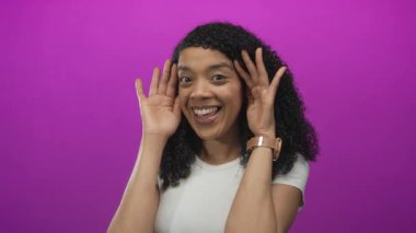 Kıvırcık saçlı bir kadın ahşap bir saat takar ve mor bir stüdyoda iki eliyle yüzünü kapatır..