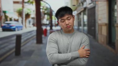 Arka planda binalar olan, gri bir kazak giyen ve kollarını kavuşturan genç Çinli bir adam açık havada neşeli bir ifade sergiliyor..