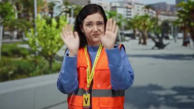 Turuncu yelekli kadın, arka planda yeşil ağaçlar olan kalabalık caddedeki gürültüden kulaklarını kapatıyor ve dışarıda rahatsızlığını dile getiriyor..