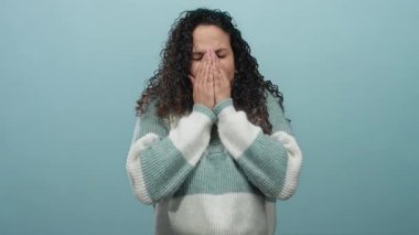 Mavi arka planda hapşıran İspanyol kadın, rahat bir kazak giyiyor ve rahatsızlığını dile getiriyor..