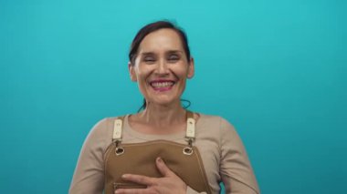 Parlak turkuaz bir duvara karşı bronz bir önlükle gülümseyen kadın neşeli bir ifadeyle dost bir atmosfer izlenimi veriyor..