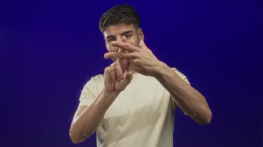 Latin kökenli genç adam mavi stüdyoda elleri ile hashtag jesti oluşturmak için parmaklarını çapraz yapıyor..
