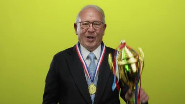 Takım elbiseli kıdemli adam, sarı arka planda zafer ve başarıyı neşeyle kutlayan kupa ve madalya taşıyor..