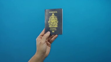 Bir adam mavi bir duvara karşı Kanada pasaportu tutuyor, izole bir ortamda seyahat kimliği gösteriyor, belgeyi ve onu tutan eli vurguluyor..
