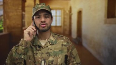 Kamuflaj üniformalı bir adam bina içinde görünür künyelerle telefonu kulağına tutuyor. Görev ve kararlılık..