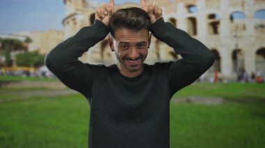 Erkekler tarihi kolezyum binasının önünde parmaklarıyla boynuz hareketi yapar ve kameraya gülümserler; neşeli seyahat anı.