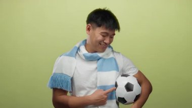 Futbol topu ve eşarpla gülümseyen genç adam izole edilmiş sarı arka plana karşı, spor için enerji ve coşku gösteriyor..