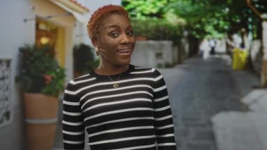 Kadın başını eğip gülümseyen yüzünü ve kısa kıvırcık saçlarını gösteriyor. Çizgili süveter giyiyor. Düşünce..