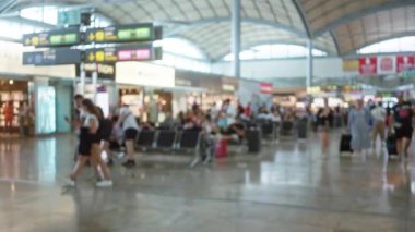 Bulanık gezginlerin yürüdüğü, beklediği ve büyük işaretler altında cihazlar kullandığı yoğun havaalanı terminali modern hava yolculuğunun hareketli ortamını yakalıyor.