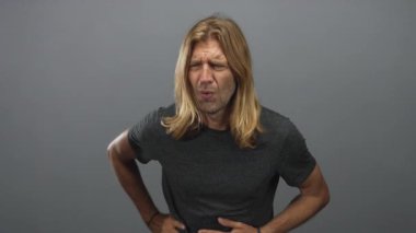 Uzun saçlı, koyu renk tişörtlü sarışın bir adam, gri stüdyo ortamında belini daha aşağıda tutar..