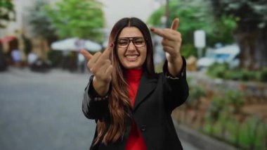 Gözlüklü, kendine güvenen genç bir kadın enerjik bir sokakta cesur bir jest yapıyor, açık havada yaramazca gülümsüyor..