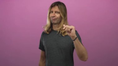 Koyu gri tişörtlü, uzun sarı saçlı bir adam stüdyo ortamında uzanmış pembe duvarlı, ezik bir jest yapıyor..