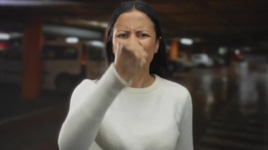 Karanlık bir otopark ortamında, beyaz bir kazak giymiş, yumruğunu sıkmış ve sinirli bir ifade sergileyen bir kadın..