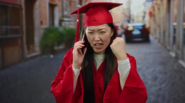Kırmızı mezuniyet elbisesi giymiş bir kadın, şehir sokak ortamında telefonla konuşurken, yumruğunu sıkarken ve kaşlarını çatarken, güçlü duygular ifade ederken hayal kırıklığına uğramış görünüyor..