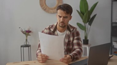Genç adam ofiste dizüstü bilgisayarla belgeyi inceliyor, endişelerini dile getiriyor, bitkilerle çevrili, odaklanmış ve ciddi ruh halini yansıtıyor..